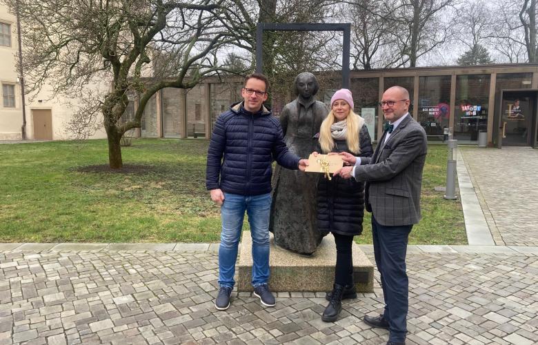 3 Menschen vor Skulptur, übergeben Briefumschlag, Bäume im Hintergrund
