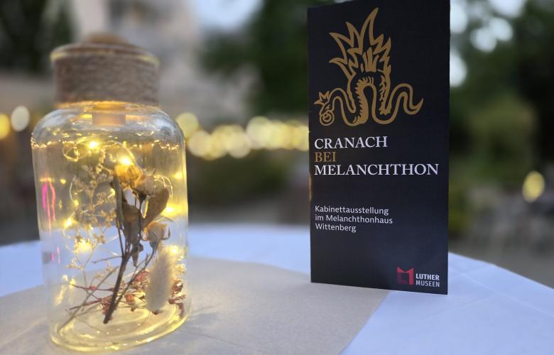 ein Licht im Glas steht auf Tisch, weiße Tischdecke, rechts daneben Hochkantkarte, grau "Cranach bei Melanchthon"