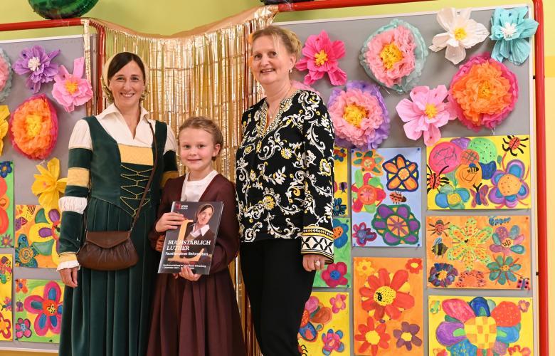2 Frauen, in der Mitte ein kleines Mädchen vor bunter Blumenwand