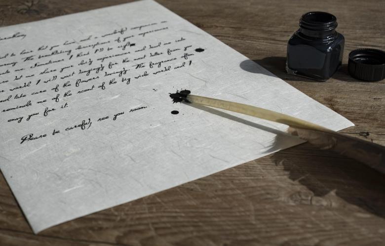 beschriebenes Papier, ein Tintenglas und eine Schreibfeder liegen auf Holztisch