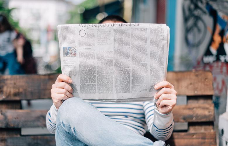ein Mensch sitzt auf einer Bank, liest Zeitung. Die Zeitung ist im Fokus
