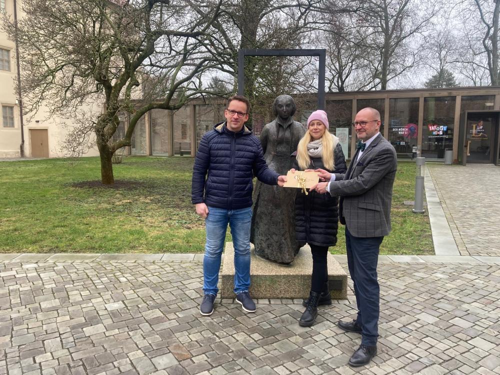 3 Menschen vor Skulptur, übergeben Briefumschlag, Bäume im Hintergrund