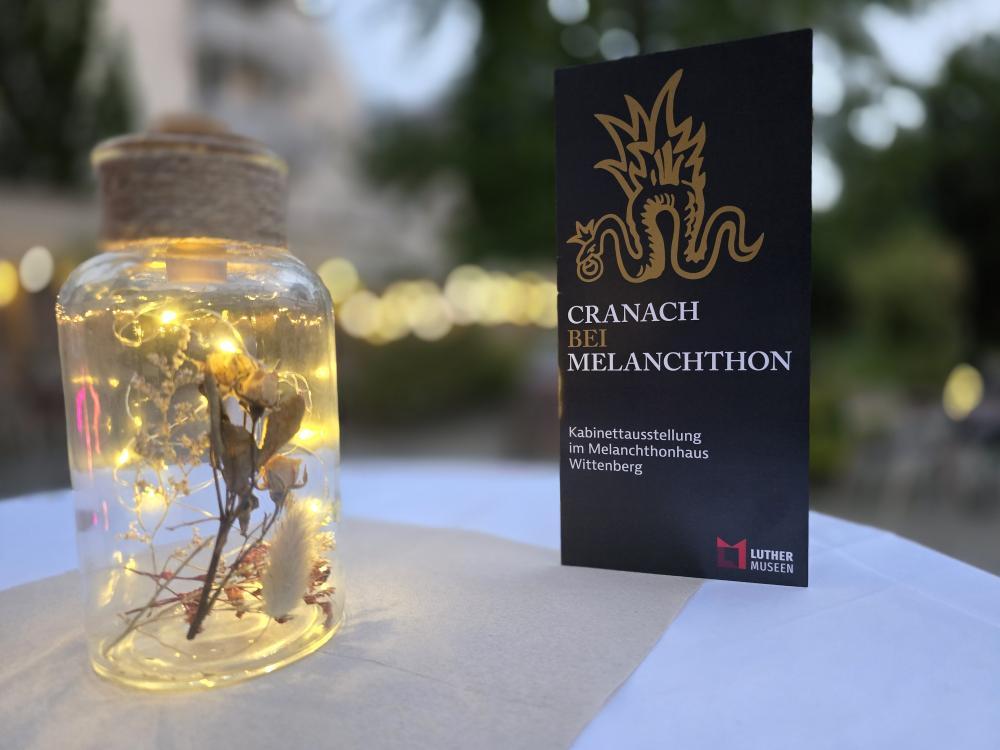 ein Licht im Glas steht auf Tisch, weiße Tischdecke, rechts daneben Hochkantkarte, grau "Cranach bei Melanchthon"