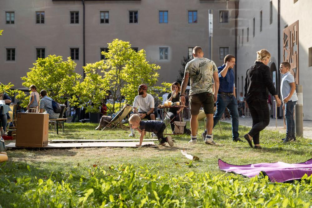Wiese, Haus im Hintergrund, Kind spielt mit Murmeln, Erwachsene sitzen und erzählen
