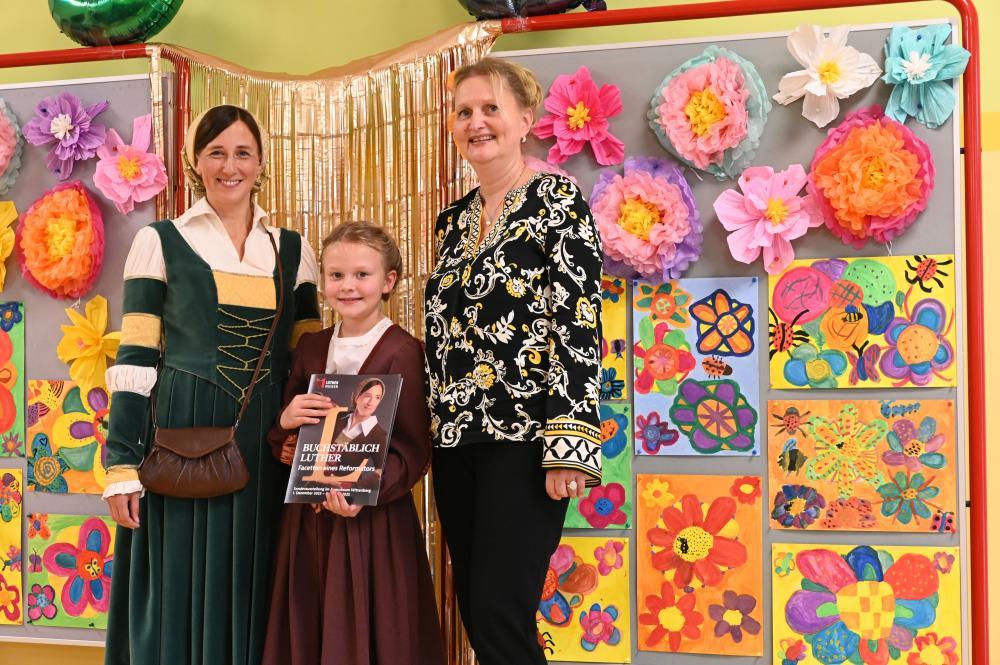 2 Frauen, in der Mitte ein kleines Mädchen vor bunter Blumenwand