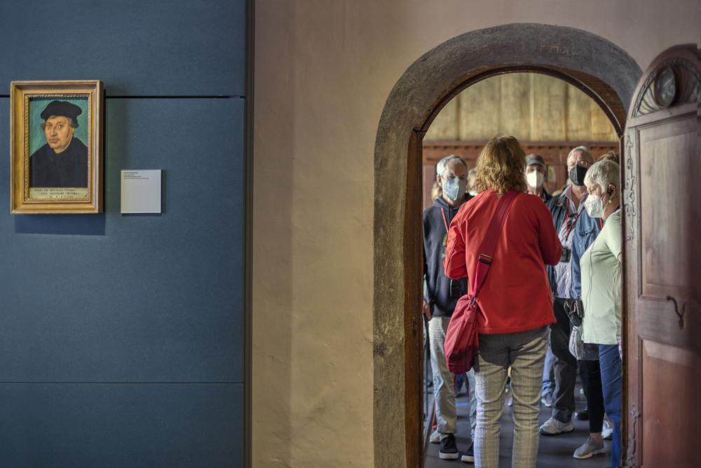 links blaue Wand mit Foto von Luther, rechts offene Tür. im nächsten Raum viele Menschen