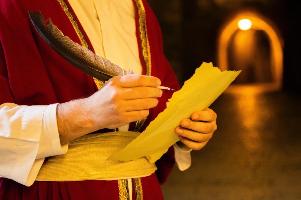 Mensch in Robe, schreibt mit Feder auf ein Stück Pergament, im Hintergrund langer schlecht beleuchteter Gang